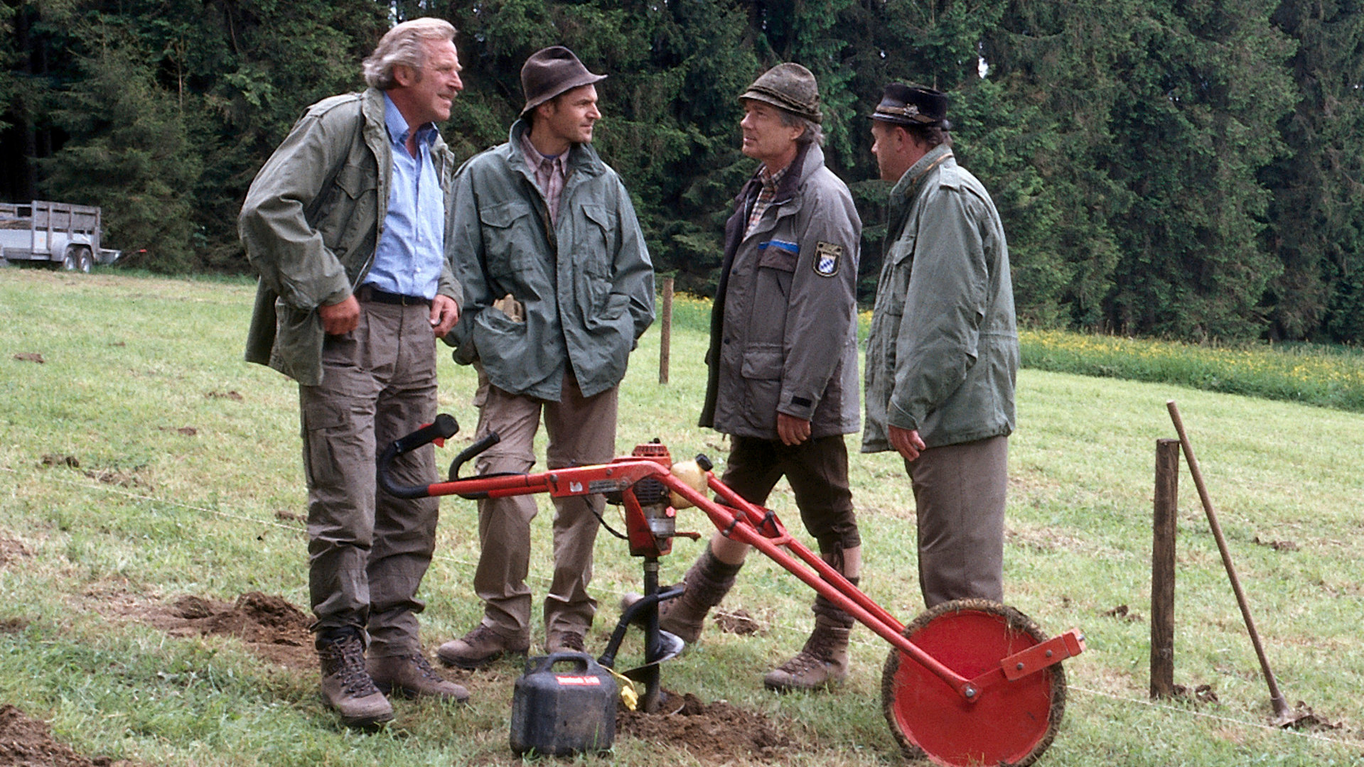 "Forsthaus Falkenau - Neue Wege": Martin Rombach (Christian Wolff) steht mit seinen Waldarbeitern (Hermann Giefer, Winfried Frey und Hans Stadlbauer) auf einer Wiese. Im Hintergrund sind Nadelbäume zu sehen. Die Männer unterhalten sich. Vor ihnen steht eine Maschine, um Löcher für die Bäume zu bohren.