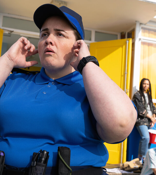 "The Outlaws - Der Protest": Diane (Jessica Gunning) hält sich die Finger an ihre Ohren und schaut etwas benebelt. Sie trägt eine blaue Uniform. Hinter ihr halten Menschen Protestschilder hoch, auf denen unter anderem "Community NOT 5G" steht.