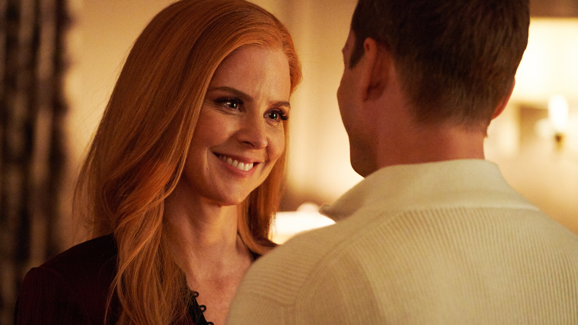 "Suits - Jetzt ist alles anders": Donna (Sarah Rafferty) steht mit langen roten Haaren vor Harvey (Gabriel Macht), dessen Rücken der Kamera zugewandt ist, und lächelt ihn strahlend und liebevoll an.