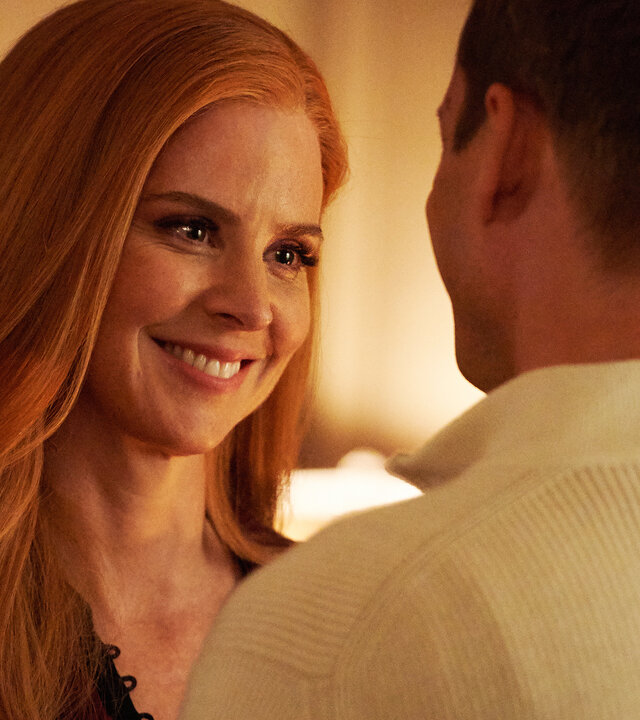 "Suits - Jetzt ist alles anders": Donna (Sarah Rafferty) steht mit langen roten Haaren vor Harvey (Gabriel Macht), dessen Rücken der Kamera zugewandt ist, und lächelt ihn strahlend und liebevoll an.