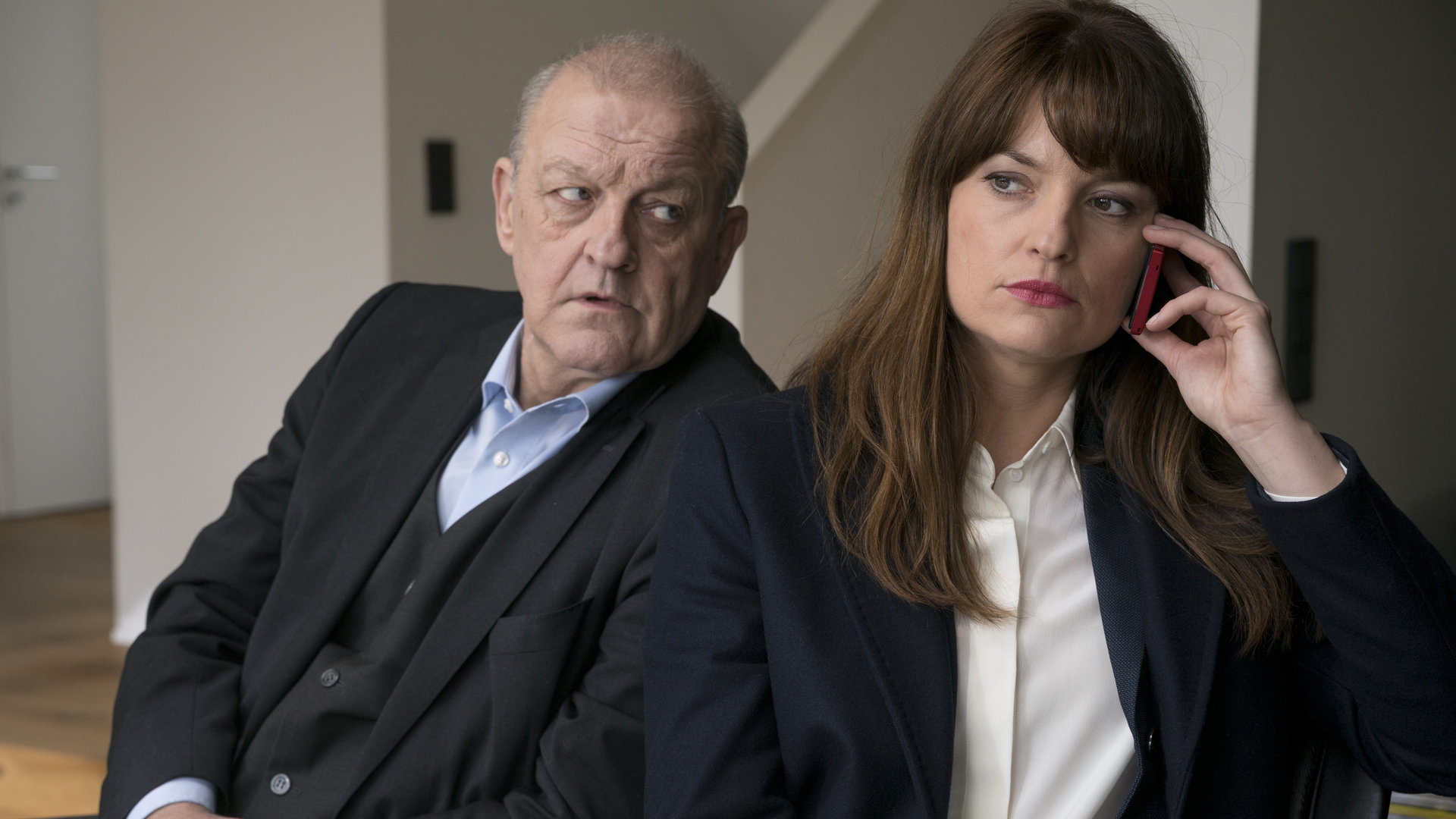 "Wilsberg - Prognose Mord": Georg Wilsberg (Leonard Lansink) und Alex Holtkamp (Ina Paule Klink)sitzen nebeneinander an einem Tisch in einer Wohnung. Alex hält ein rotes Handy am Ohr und schaut nach vorne. Wilsberg blickt sehr skeptisch und fragend zu ihr rüber.