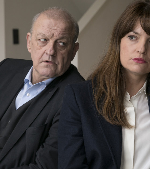 "Wilsberg - Prognose Mord": Georg Wilsberg (Leonard Lansink) und Alex Holtkamp (Ina Paule Klink)sitzen nebeneinander an einem Tisch in einer Wohnung. Alex hält ein rotes Handy am Ohr und schaut nach vorne. Wilsberg blickt sehr skeptisch und fragend zu ihr rüber.