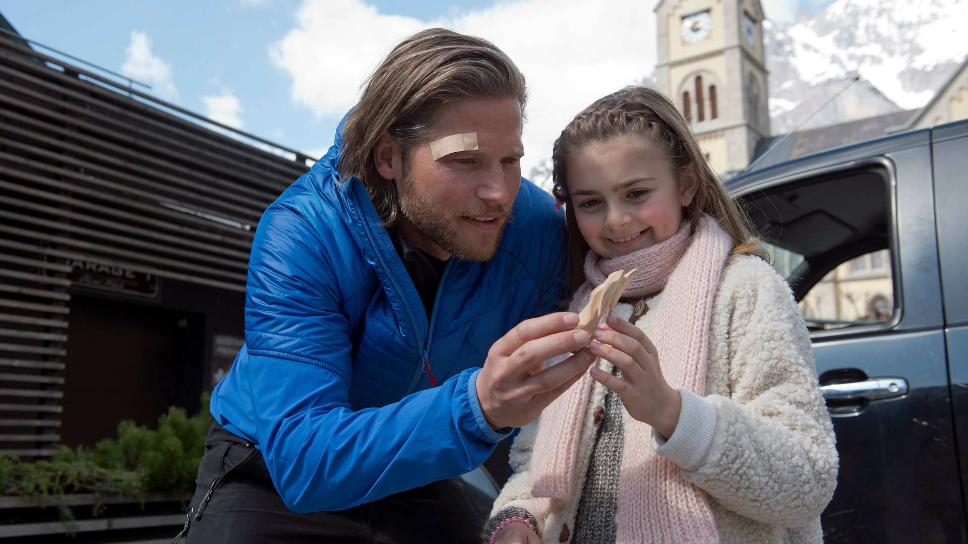 "Die Bergretter: Entscheidung im Eis": Markus (Sebastian Ströbel) hat sich zu Mia (Mia-Sophie Ballauf) gebeugt und überreicht ihr einen geschnitzten Holzengel. Mia lacht. Markus hat ein Pflaster über der rechten Augenbraue. Im Hintergrund steht ein Auto.