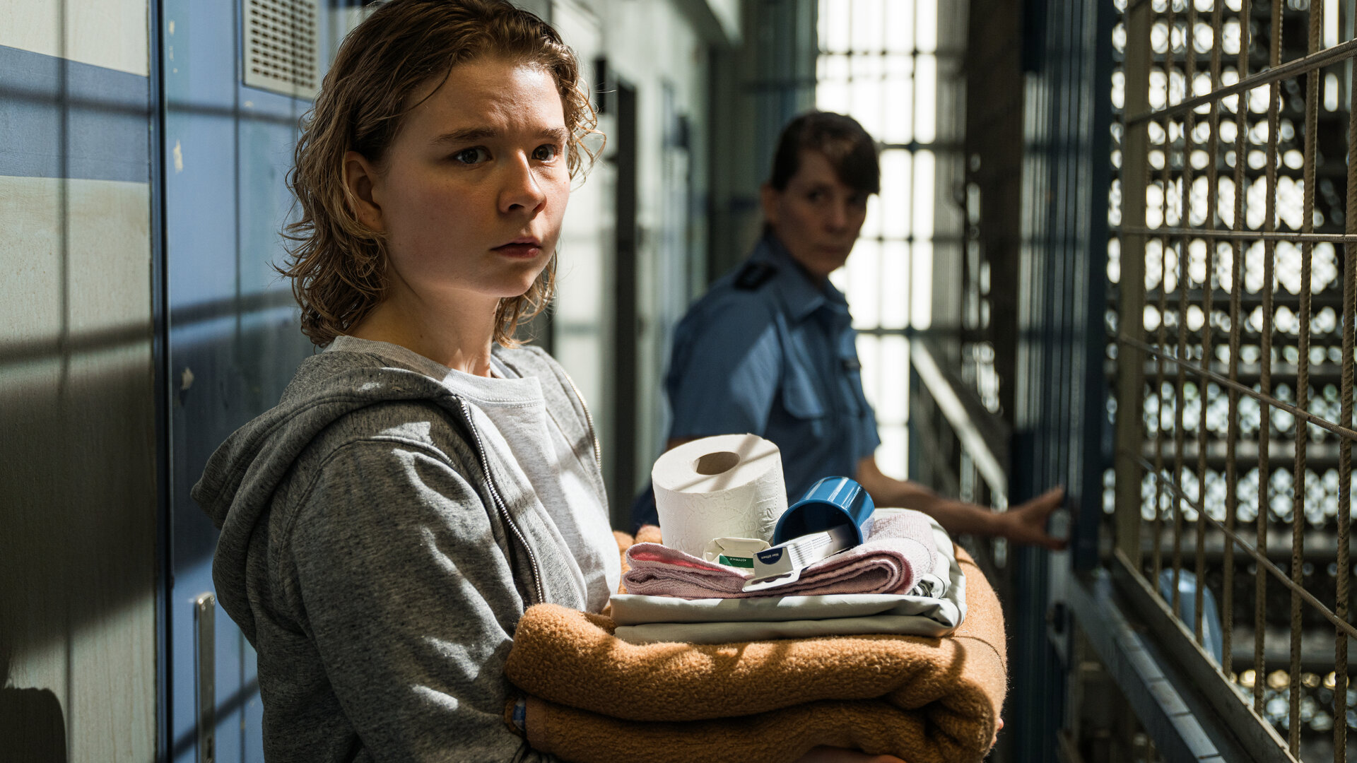 "Ein Fall für zwei - Schuld und Sühne": Mina Wirtz (Daria Vivien Wolf) steht mit besorgtem Blick auf einem Gefängniskorridor. Sie trägt einen grauen Kapuzenpullover und hält persönliche Gegenstände in den Armen. Eine Justizvollzugsbeamtin (Komparsin) steht hinter ihr.
