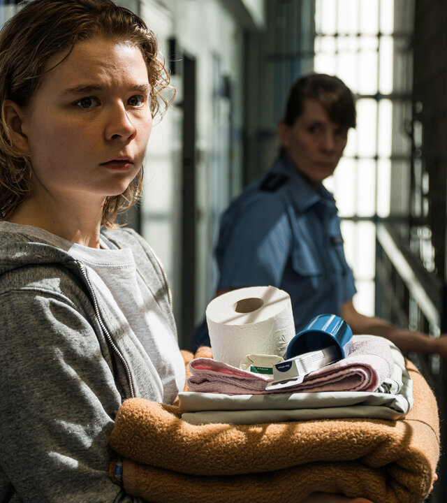 "Ein Fall für zwei - Schuld und Sühne": Mina Wirtz (Daria Vivien Wolf) steht mit besorgtem Blick auf einem Gefängniskorridor. Sie trägt einen grauen Kapuzenpullover und hält persönliche Gegenstände in den Armen. Eine Justizvollzugsbeamtin (Komparsin) steht hinter ihr.