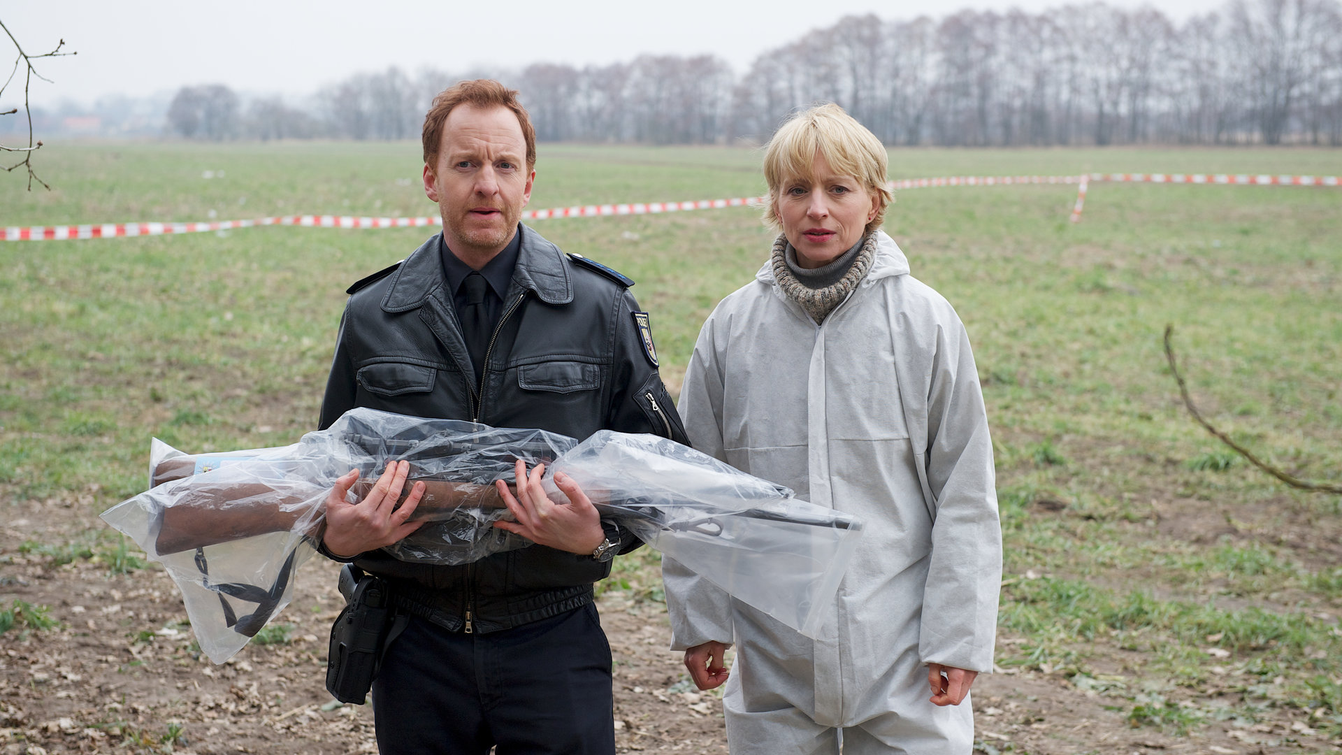 "SOKO Wismar - Tod im Feld": Kai Timmermann (Mathias Junge) und Roswitha Prinzler (Silke Matthias) stehen auf einem Feld. Timmermann hält ein Gewehr vor sich, das in Schutzfolie eingewickelt ist.