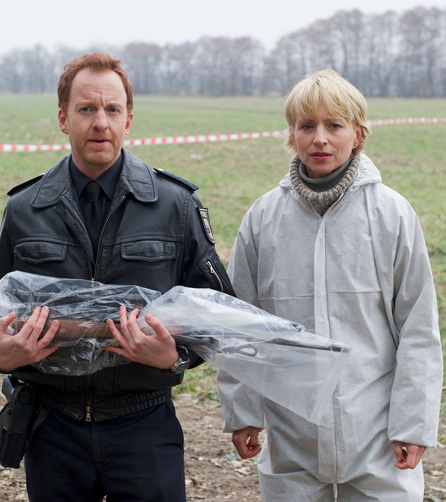 "SOKO Wismar - Tod im Feld": Kai Timmermann (Mathias Junge) und Roswitha Prinzler (Silke Matthias) stehen auf einem Feld. Timmermann hält ein Gewehr vor sich, das in Schutzfolie eingewickelt ist.