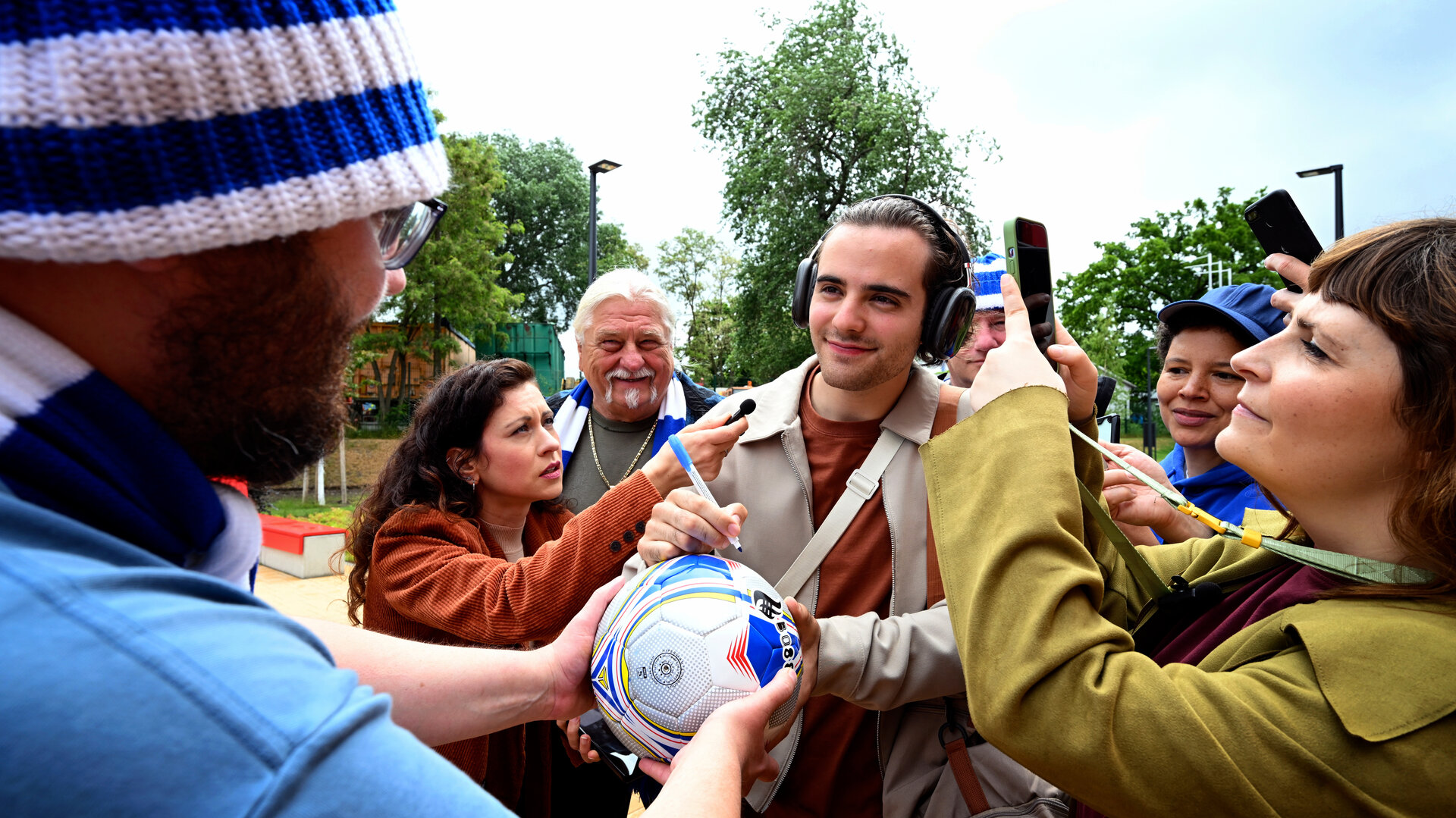 "Jenseits der Spree - Abseits": László Varga (Christoph Moreno) wird auf dem Fußballplatz von Journalisten und Fans umringt und gibt gerade ein Autogramm auf einen Fußball.