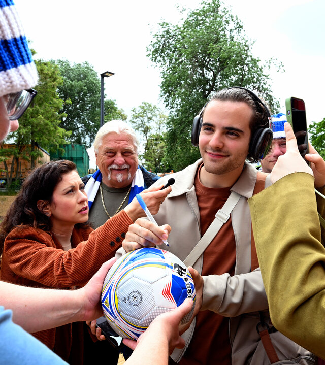 "Jenseits der Spree - Abseits": László Varga (Christoph Moreno) wird auf dem Fußballplatz von Journalisten und Fans umringt und gibt gerade ein Autogramm auf einen Fußball.