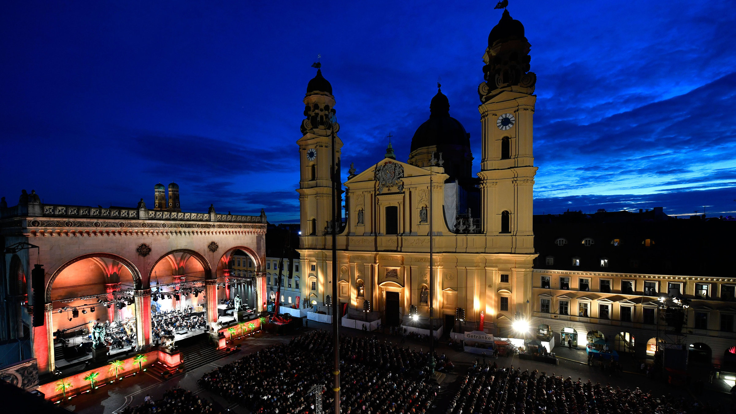 "Klassik am Odeonsplatz: Yuja Wang": Der Münchner Odeonsplatz in der Abenddämmerung mit sitzendem Publikum. Theatinerkirche und Feldherrnhalle sind beleuchtet. In der Feldherrnhalle sitzen die Münchner Philharmoniker.