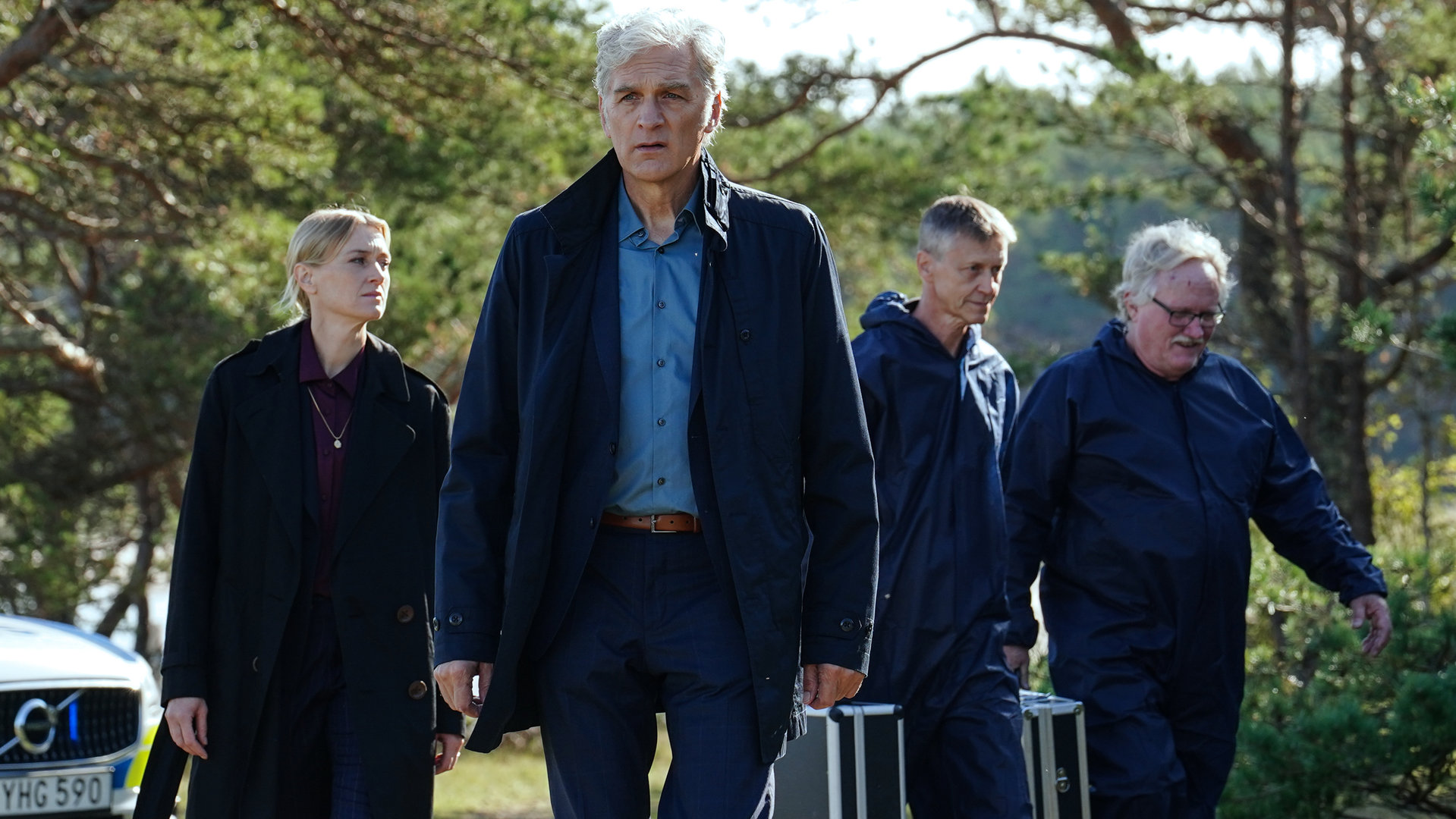 "Der Kommissar und das Meer - Woher wir kommen, wohin wir gehen": Anna-Maja Haglund (Maria Alm Norell) und Kommissar Robert Anders (Walter Sittler) schreiten durch einen offenen, stark besonnten Wald. Begleitet werden sie von zwei Mitarbeitern des forensischen Teams, die beide einen Koffer tragen. Am Bildrand ist ein Polizeiwagen zu sehen.