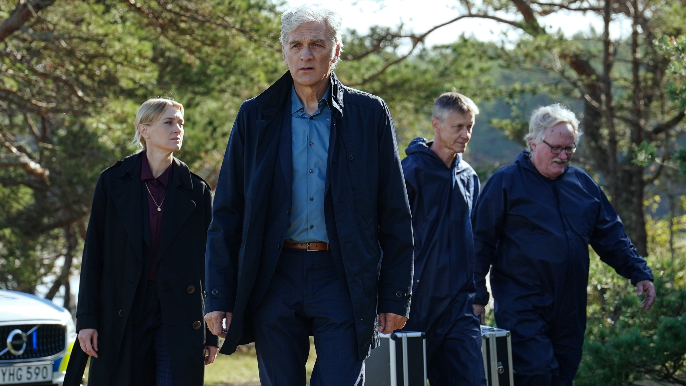"Der Kommissar und das Meer - Woher wir kommen, wohin wir gehen": Anna-Maja Haglund (Maria Alm Norell) und Kommissar Robert Anders (Walter Sittler) schreiten durch einen offenen, stark besonnten Wald. Begleitet werden sie von zwei Mitarbeitern des forensischen Teams, die beide einen Koffer tragen. Am Bildrand ist ein Polizeiwagen zu sehen.