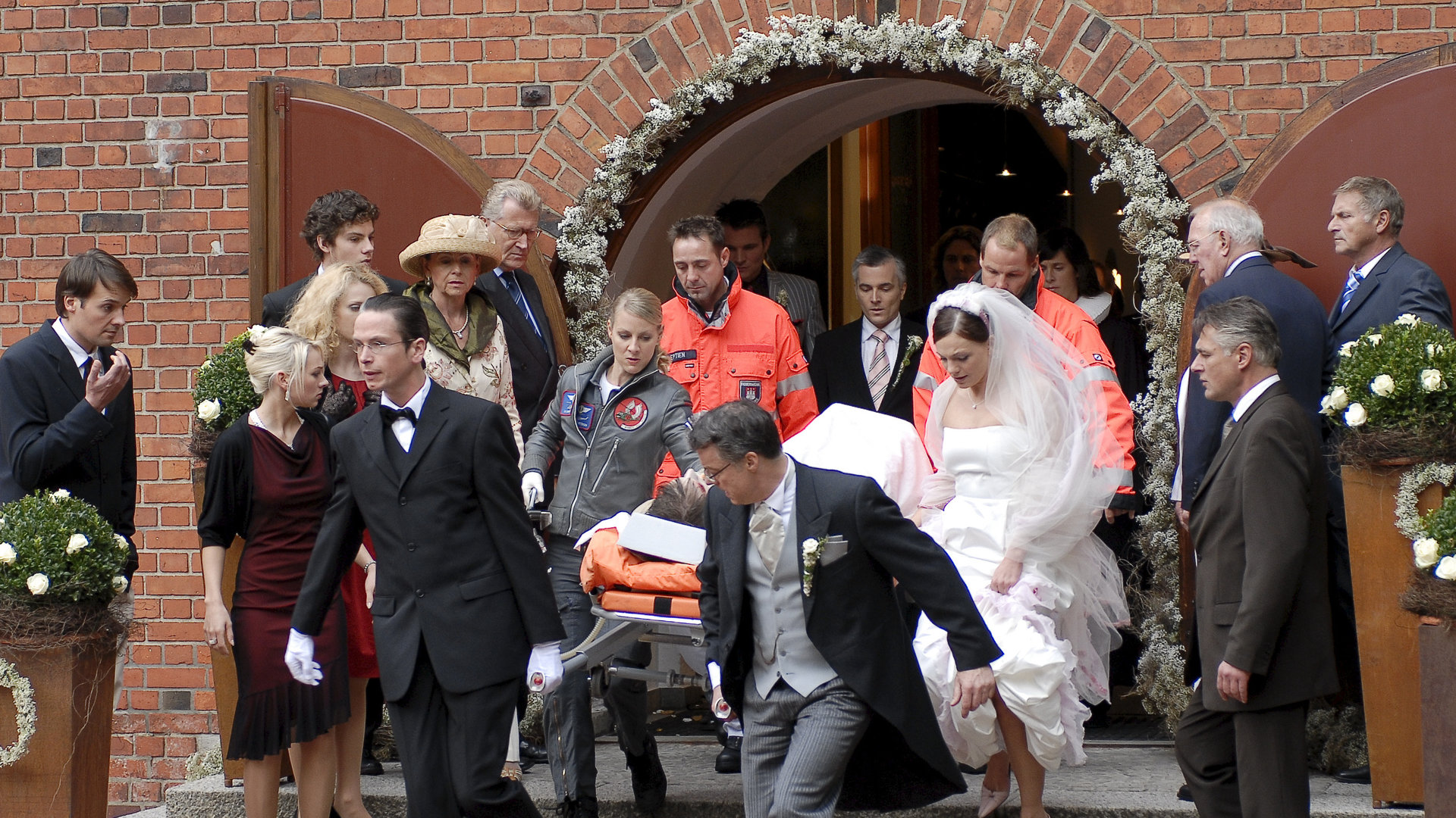 "Die Rettungsflieger - Für immer und ewig": Das Team der Rettungsflieger trägt bei einer Hochzeit eine Person aus der Kirche. Drum herum stehen die Hochzeitsgäste und die Braut Patricia Falkenstein (Anna Bertheau) läuft mit gerafften Röcken neben der Trage her.