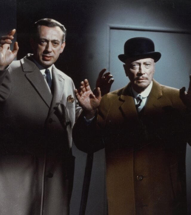 "Edgar Wallace: Der Mann mit dem Glasauge": Inspektor Perkins (Horst Tappert) und Sir Arthur (Hubert von Meyerinck) stehen mit erhobenen Händen nebeneinander in einem düsteren Raum und blicken ernst, während eine Waffe bedrohlich auf sie gerichtet ist.