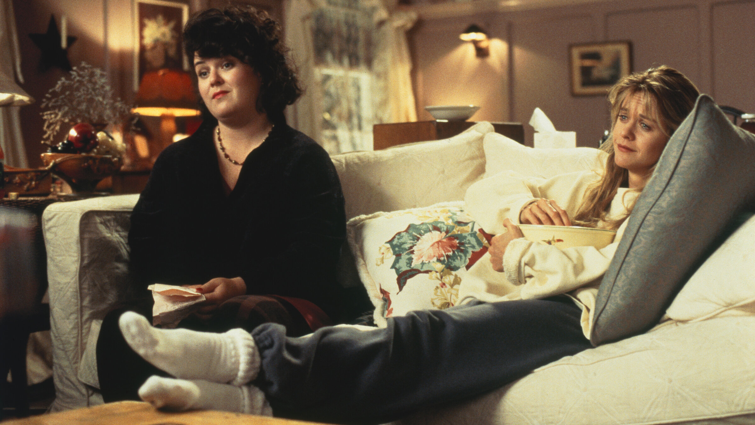 "Schlaflos in Seattle": Annie (Meg Ryan) und Becky (Rosie O'Donnell) sitzen gemeinsam auf einem Sofa im Wohnzimmer und blicken träumerisch und nachdenklich geradeaus. Dabei hält Becky mehrere Papierzettel in ihrer Hand.