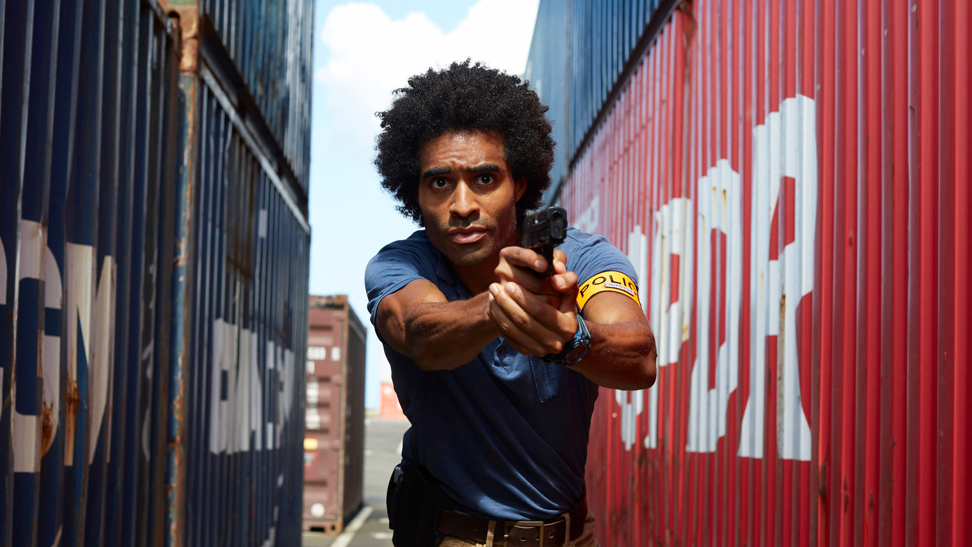 "Deadly Tropics - Ein Sohn vergisst nie": Aurélien Charlery (Julien Béramis) rennt zwischen Containern entlang und zielt mit seiner Waffe, die er in seinen Händen hält. Sein Blick ist fokussiert in die Ferne gerichtet.