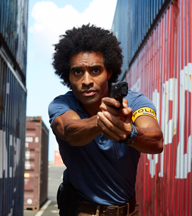 "Deadly Tropics - Ein Sohn vergisst nie": Aurélien Charlery (Julien Béramis) rennt zwischen Containern entlang und zielt mit seiner Waffe, die er in seinen Händen hält. Sein Blick ist fokussiert in die Ferne gerichtet.