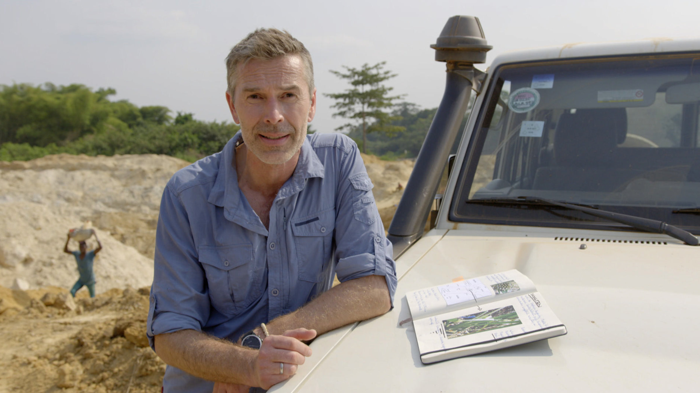 "Terra X: Rätselhafte Phänomene - Diamantenfieber und sprechende Pflanzen": Dirk Steffens lehnt an einem Jeep und schaut in die Kamera. Auf der Kühlerhaube liegt ein Notizbuch mit dem Foto einer Pflanze. Im Hintergrund ist ein Steinbruch zu sehen, in dem ein Mann steht, der einen Felsbrocken über dem Kopf trägt.