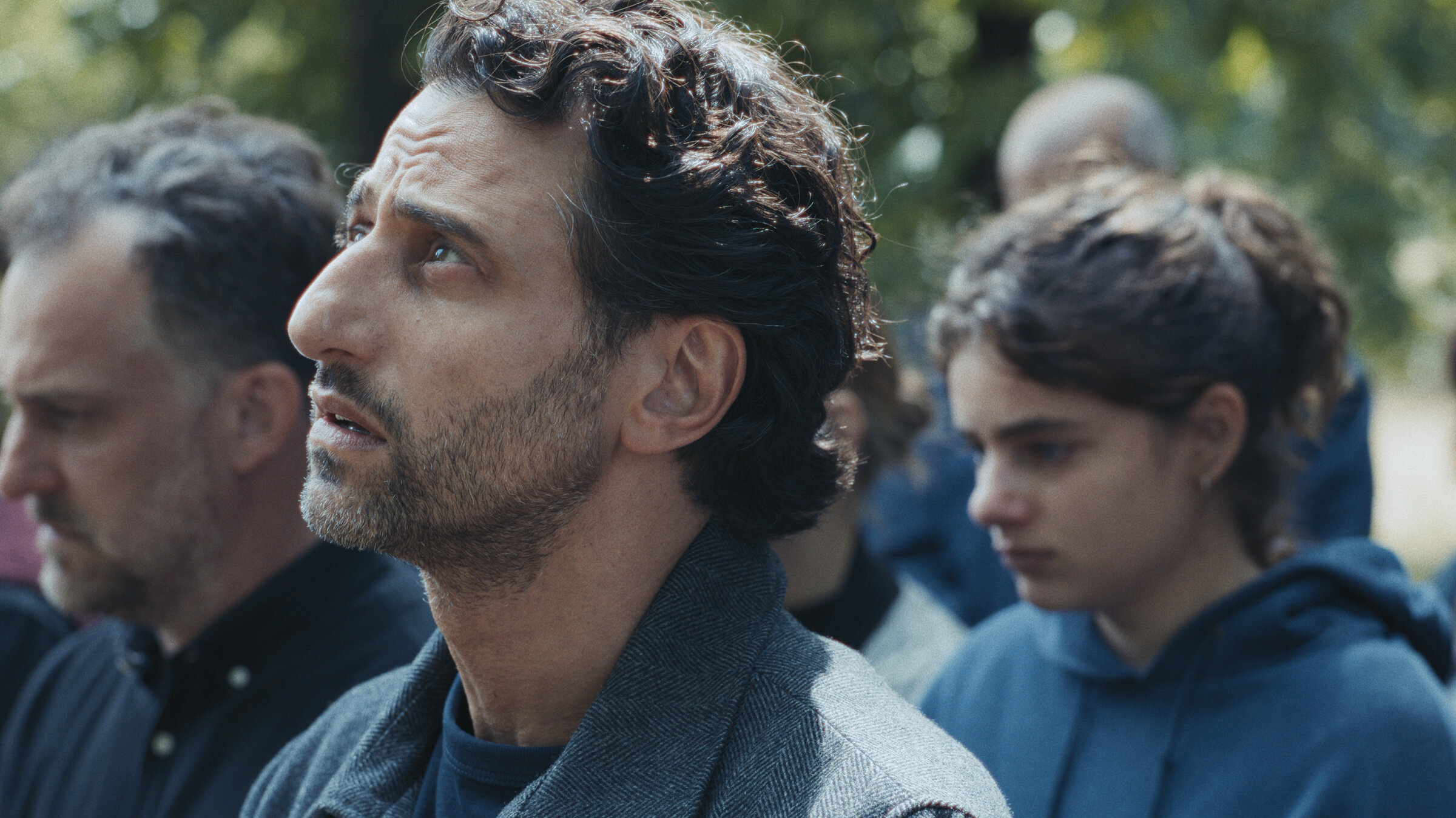 „Was von der Liebe bleibt“: Zwischen einer Gruppe von Menschen steht Ilyas (Serkan Kaya), dessen Blick suchend nach oben gerichtet ist. Seine Tochter Senna (Amira Demirkiran) senkt daneben den Kopf, als wollte sie sich zurückziehen. Beide wirken tief betroffen.