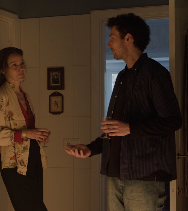 "This Is Gonna Be Great - Familie": David (Rein Mulder) steht in einem Raum vor einer geöffneten Tür. Neben ihm lehnt Monika (Jeanette Hain) an einem Schrank. Die beiden sprechen miteinander.