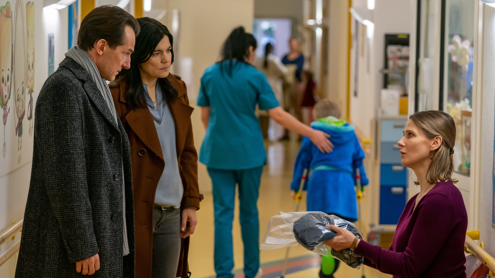 "Letzte Spur Berlin - Sorgenkind": Radek (Hans-Werner Meyer) und Mina (Jasmin Tabatabai) stehen in einem Krankenhausflur. Ihnen gegenüber sitzt Janina (Katrin Röver), die den Schlafanzug ihres Sohnes entgegennimmt. Sie schaut sehr besorgt zu Radek und Mina auf.