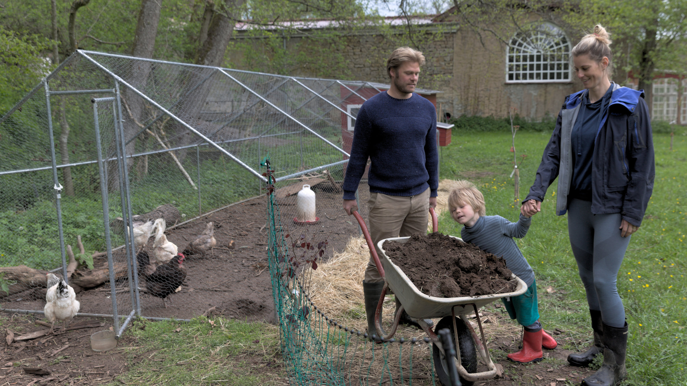 "37° Unsere eigene Farm - Selbstversorgen lernen": Michael und Sara stehen ihrem kleinen Sohn Max und einer Schubkarre, gefüllt mit Erde, neben dem Außengehege eines Hühnerstalls. Man sieht auch weiße und braune Hühner.