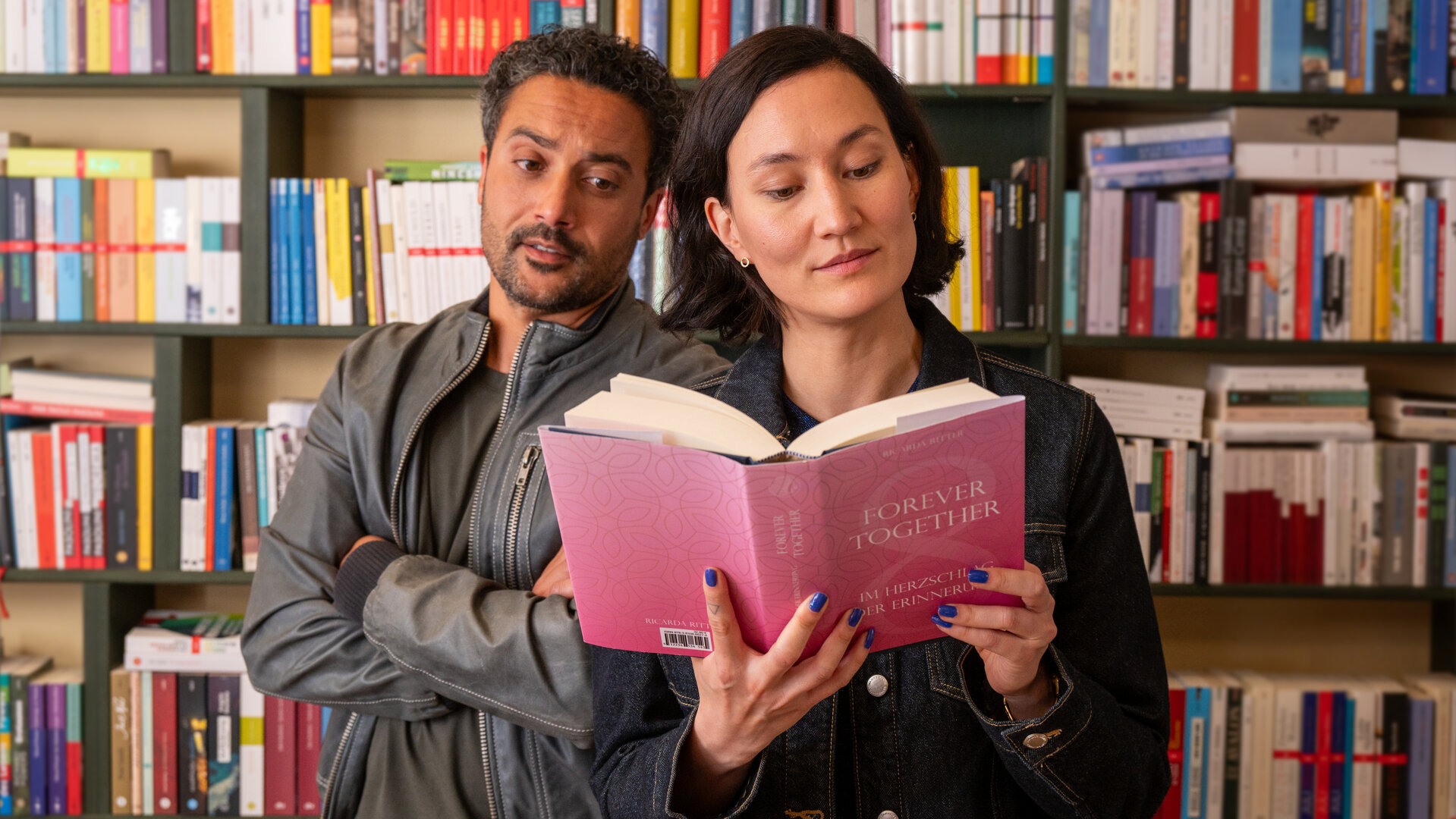 "SOKO Potsdam - Forever Together": In einem hellen Raum einer Buchhandlung stehen David Grünbaum (Omar El-Saeidi) und Pauline Hobrecht (Agnes Decker) dicht nebeneinander vor einem Regal voller Bücher. Pauline hält ein rosafarbenes Buch mit dem Titel "Forever Together – Im Herzschlag der Erinnerung" in den Händen und liest konzentriert. David steht leicht hinter ihr, verschränkt die Arme und blickt mit nachdenklicher Miene auf das Buch. Hinter ihnen reihen sich bunt sortierte Buchrücken aneinander.