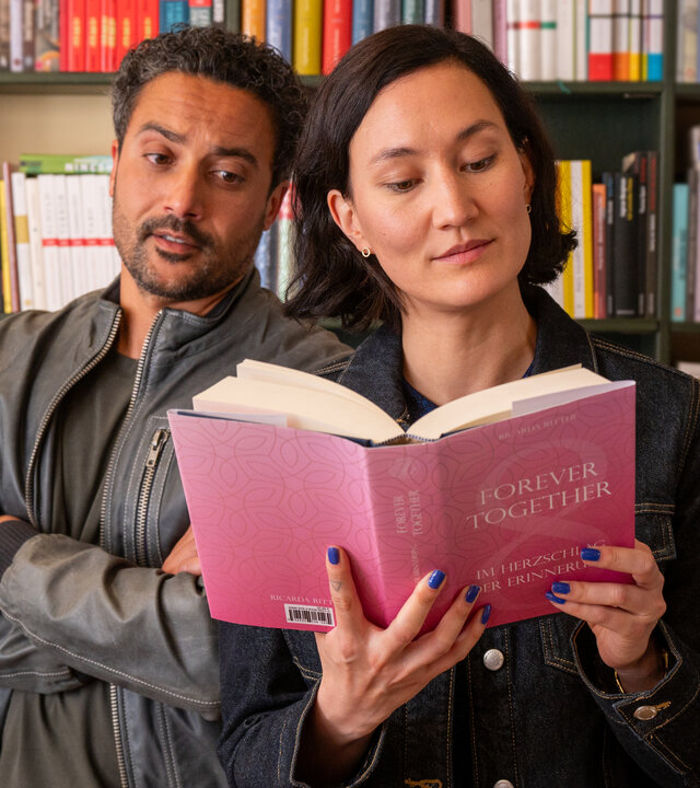 "SOKO Potsdam - Forever Together": In einem hellen Raum einer Buchhandlung stehen David Grünbaum (Omar El-Saeidi) und Pauline Hobrecht (Agnes Decker) dicht nebeneinander vor einem Regal voller Bücher. Pauline hält ein rosafarbenes Buch mit dem Titel "Forever Together – Im Herzschlag der Erinnerung" in den Händen und liest konzentriert. David steht leicht hinter ihr, verschränkt die Arme und blickt mit nachdenklicher Miene auf das Buch. Hinter ihnen reihen sich bunt sortierte Buchrücken aneinander.