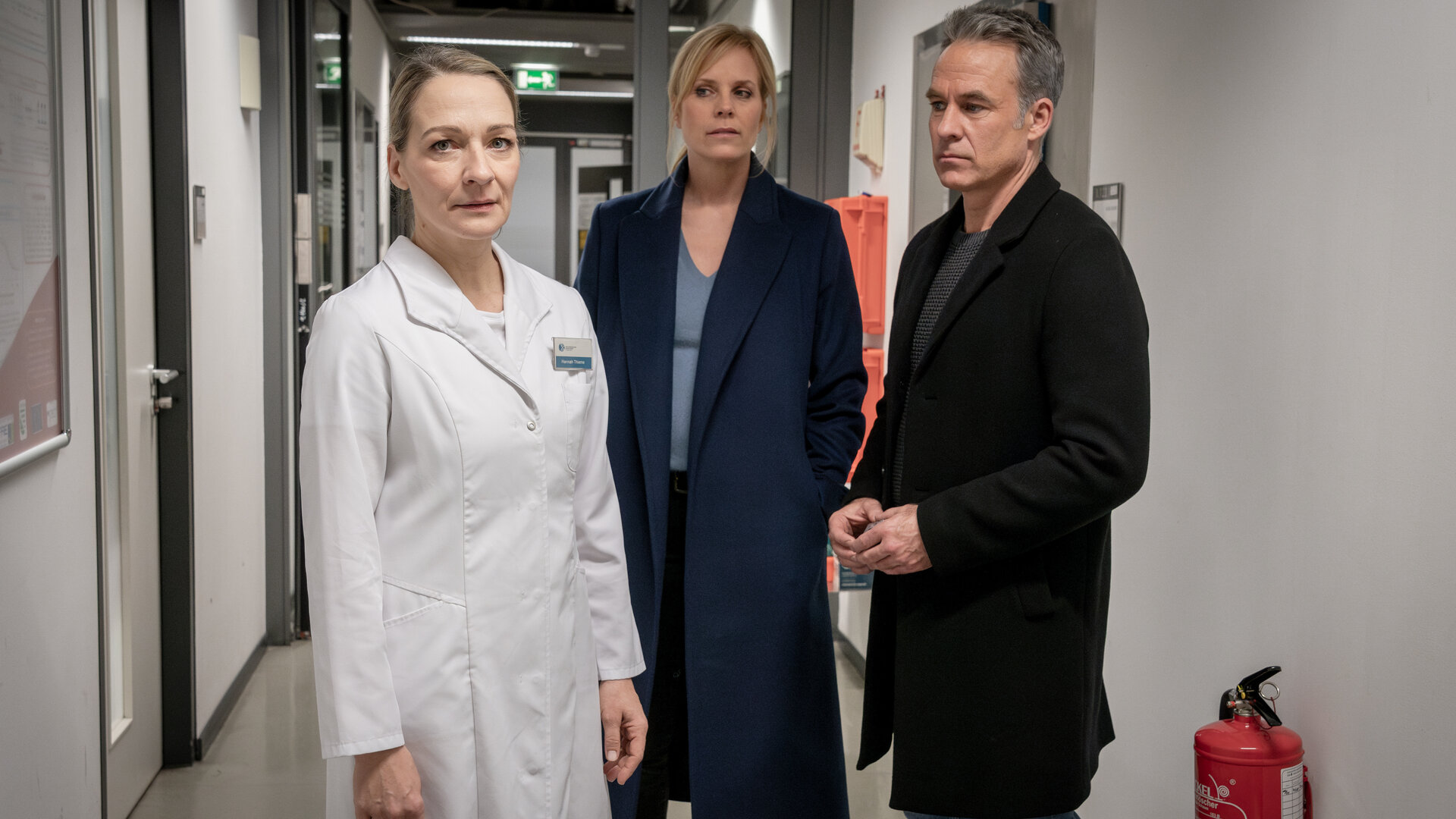 "SOKO Leipzig - Um ein Haar": Ina (Melanie Marschke) und Jan (Marco Girnth) stehen in einem Gang, der sehr klinisch wirkt. Sie schauen zu Laborassistentin Hannah Thieme (Anika Mauer) an, die neben ihnen steht und in die Kamera schaut. Sie trägt einen weißen Kittel mit ihrem Namensschild.