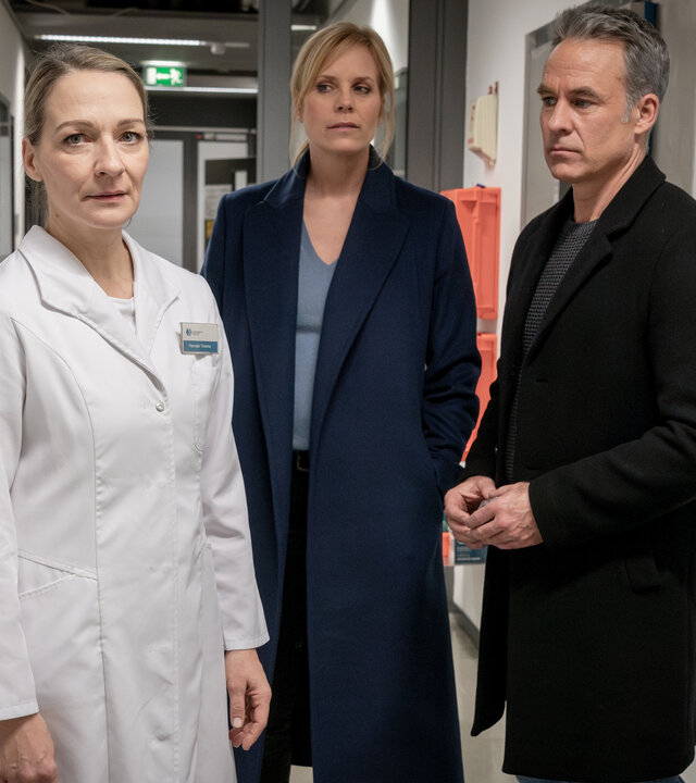"SOKO Leipzig - Um ein Haar": Ina (Melanie Marschke) und Jan (Marco Girnth) stehen in einem Gang, der sehr klinisch wirkt. Sie schauen zu Laborassistentin Hannah Thieme (Anika Mauer) an, die neben ihnen steht und in die Kamera schaut. Sie trägt einen weißen Kittel mit ihrem Namensschild.
