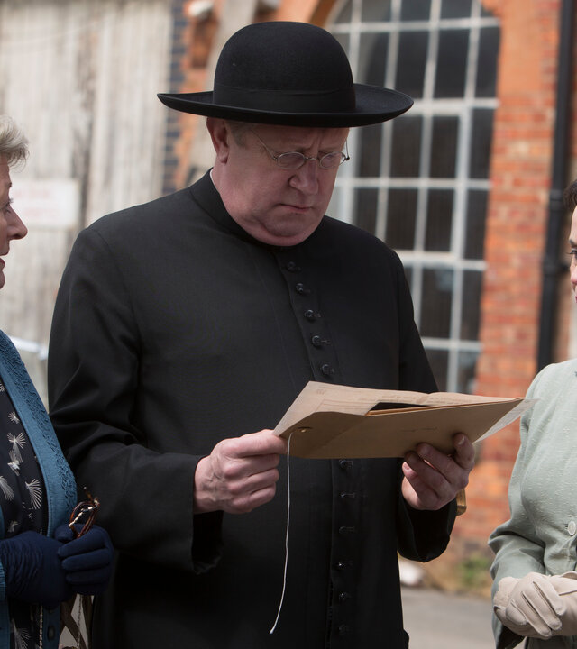 "Father Brown - Der Narr der Nation": Mrs. McCarthy (Sorcha Cusack), Father Brown (Mark Williams) und Annabelle Marcel (Oona Kirsch) stehen nebeneinander vor einem Gebäude. Father Brown liest einen Brief, die beiden Damen wirken besorgt.