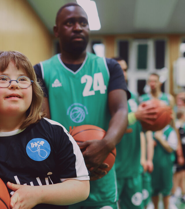 "Aktion Mensch - Glückszahlen der Woche": Mehrere Personen stehen in einer Sporthalle. Im Vordergrund steht ein Mädchen neben einem Mann. Beide schauen lächelnd zur Seite.