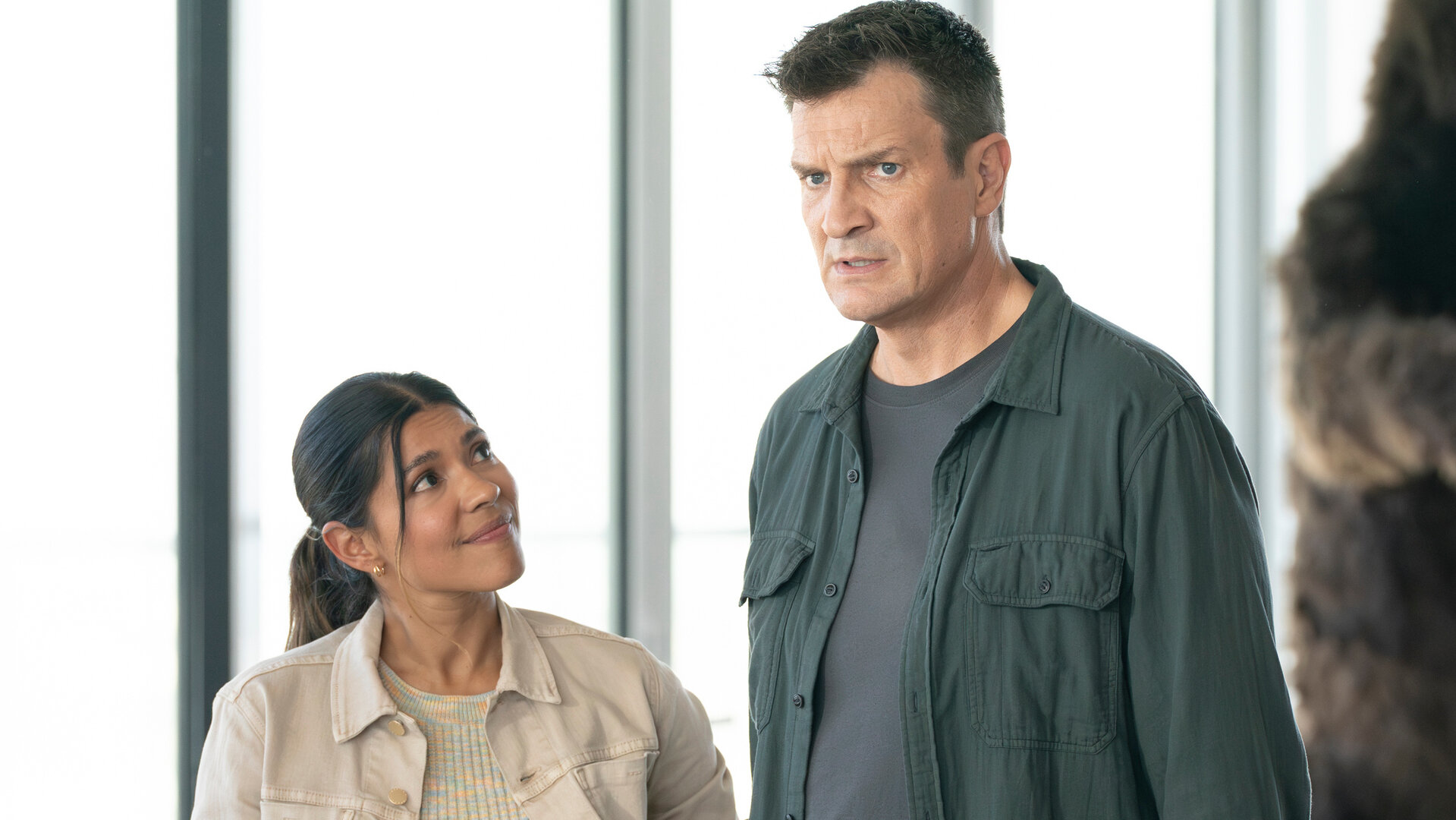 "The Rookie - Grumpy Cop": Officer John Nolan (Nathan Fillion) und Celina Juarez (Lisseth Chavez) stehen nebeneinander vor einer großen Fensterfront. Beide tragen Zivilkleidung. Juarez blickt mit einem erwartungsvollen Ausdruck zu Nolan, der mit einem ernsten Ausdruck nach vorne blickt.
