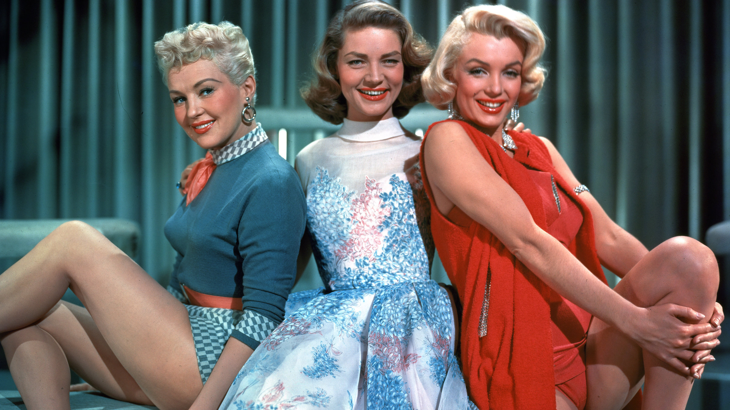 "Wie angelt man sich einen Millionär?": Drei junge Frauen, die den 1950erJahren entsprechende modische Sommerkleidung tragen, posieren auf grünen Hockern und blicken keck in die Kamera. Die linke trägt kurzes blondes Haar, Kreolen und ein grünes Oberteil mit passenden Shorts, die Mittlere schulterlanges brünettes Haar und ein geblümtes Sommerkleid, die rechte wasserstoffblondes Haar und einen roten Badeanzug mit passender Weste.