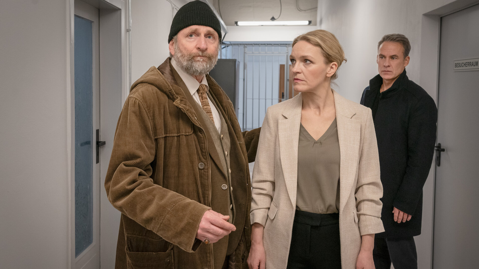 "SOKO Leipzig - Angeklagt": Jan (Marco Girnth), Leni (Caroline Scholze) und Dr. Egon Fleischbauer (Jörg Witte) stehen in einem grauen Flur. Im Hintergrund sind Gitterstäbe zu sehen. Fleischbauer steht neben Leni, die ihn anschaut. Er drückt seine Hand gegen Lenis Rücken und schaut nach vorne. Hinter Leni steht Jan und schaut Fleischbauer an.
