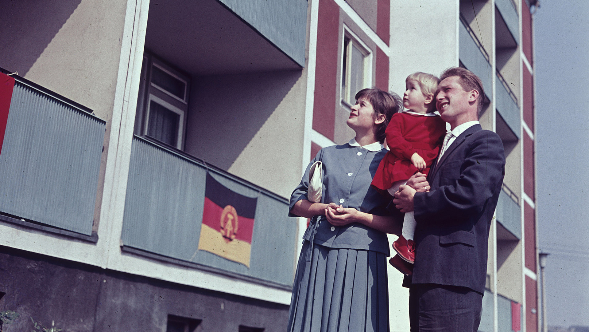 "ZDF-History: Mythen der Gegenwart – Von der Platte bis zur Autobahn": Eine junge Frau und ein junger Mann, der ein kleines Mädchen auf dem Arm hält, stehen vor einem Plattenbau in Ostberlin 1963. Sie schauen an der Fassade nach oben, die mit DDR-Fahnen geschmückt ist.