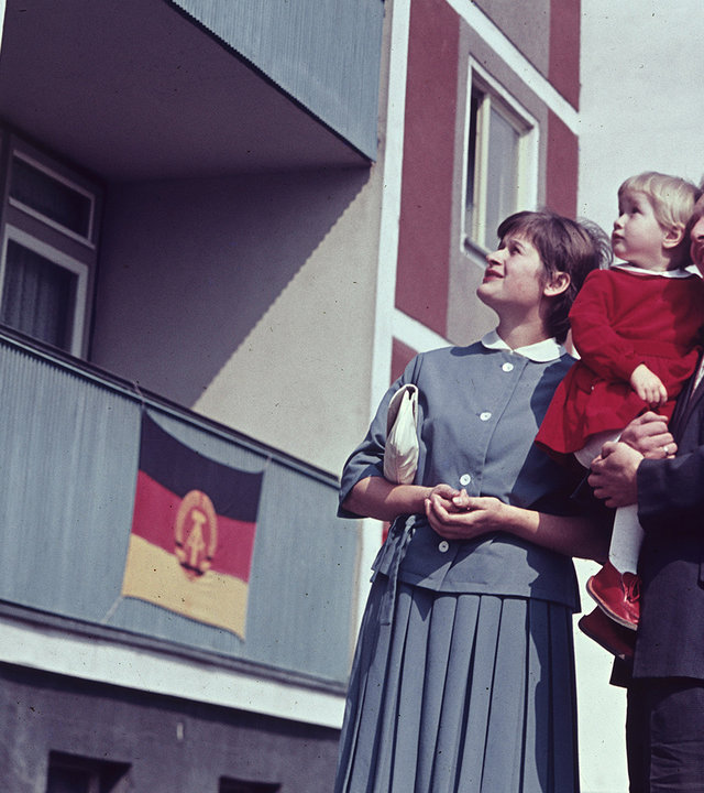"ZDF-History: Mythen der Gegenwart – Von der Platte bis zur Autobahn": Eine junge Frau und ein junger Mann, der ein kleines Mädchen auf dem Arm hält, stehen vor einem Plattenbau in Ostberlin 1963. Sie schauen an der Fassade nach oben, die mit DDR-Fahnen geschmückt ist.