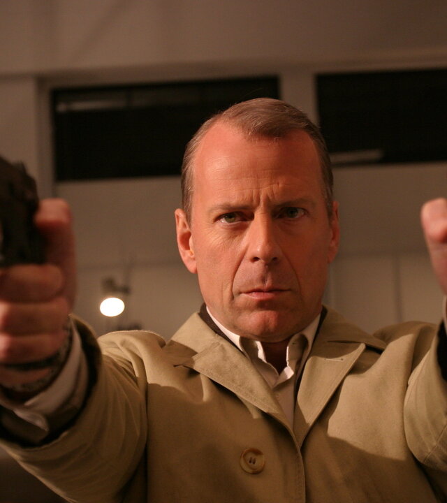"Lucky Number Slevin": Mr. Goodkat (Bruce Willis) steht in einem Wohnzimmer und hält mit ausgestreckten Armen zwei Pistolen vor sich, die in den Raum gerichtet sind. Sein Blick ist dabei fokussiert und entschlossen.