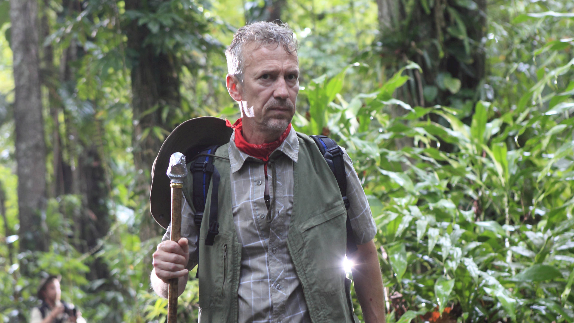 "Death in Paradise - Der Ruf der Wildnis": Ornithologe Marc Talbot (Richard Huw) steht im Dschungel. In der rechten Hand hält er einen großen Stock, auf dem er sich abstützt. Er ist in Tarnfarben gekleidet, um den Hals trägt er ein rotes Tuch. Sein Hut hängt an einer Schnur auf dem Rücken. Rundherum große Dschungelpflanzen und Bäume.
