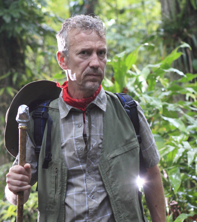 "Death in Paradise - Der Ruf der Wildnis": Ornithologe Marc Talbot (Richard Huw) steht im Dschungel. In der rechten Hand hält er einen großen Stock, auf dem er sich abstützt. Er ist in Tarnfarben gekleidet, um den Hals trägt er ein rotes Tuch. Sein Hut hängt an einer Schnur auf dem Rücken. Rundherum große Dschungelpflanzen und Bäume.