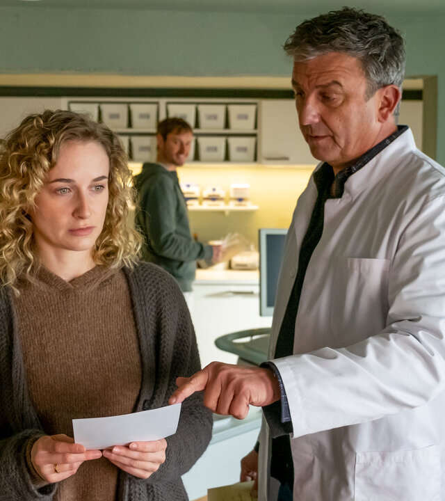 "Der Bergdoktor: Sternenkinder": In der Arztpraxis: Nachdenklich hält Jana Weiss (Pia Barucki) ein Foto in den Händen. Dr. Martin Gruber (Hans Sigl), der neben ihr steht, zeigt mit dem Finger darauf. Im Hintergrund steht David Kästner (Frédéric Brossier) und beobachtet die beiden.