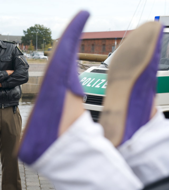 "SOKO Wismar - Spieglein, Spieglein": Leena Virtanen (Li Hagman) und Lars Pöhlmann (Dominic Boeer) stehen mit ihrem Polizeiauto am Hafen. Vor ihnen sind zwei Füße in lilafarbenen Schuhen zu sehen, die mit weißen Hosen über den Knöcheln aus einem schwarzen Müllcontainer herausragen.