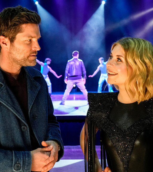 "SOKO Stuttgart - Falsche Töne": Kriminalhauptkommissar Jo Stoll (Peter Ketnath) befragt in einer Veranstaltungshalle die Schlagersängerin Verena Herz (Sarah Thonig). Im Hintergrund proben gerade die Tänzer mit einem Choreografen (Eric Gauthier) auf einer Bühne.