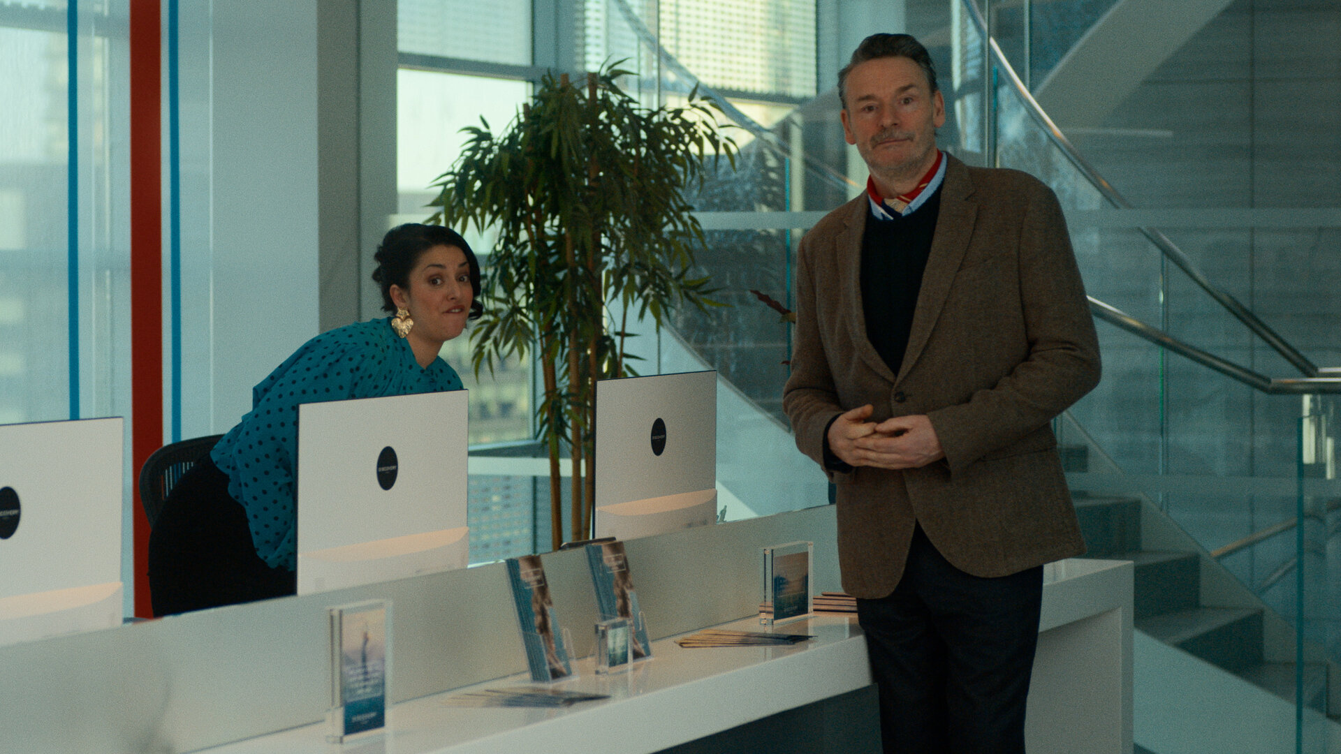 "Extraordinary - Auf Nimmerwiedersehen": Georges Assistentin ( Mona Goodwin) sitzt fast stehend im feinem Blüschen hinter der weißen Kliniktheke und verzieht ihr Gesicht. Psychologe George (Julian Barratt) steht mit verschränkten Händen vor der Theke und schaut unschuldig drein.