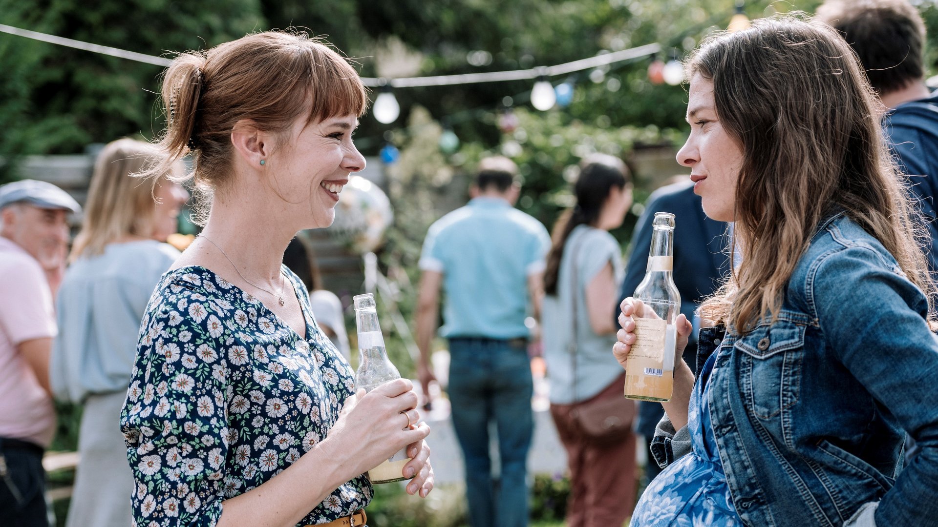 "DEUTSCHER: Folge 3 - Was morgen ist": Ulrike Pielcke (Milena Dreißig) und Eva Schneider (Meike Droste) stehen sich beim Gartenfest gegenüber. Beide halten Saftflaschen in den Händen. Eva Schneider ist hochschwanger.