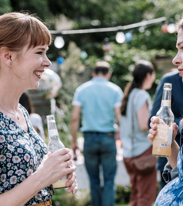 "DEUTSCHER: Folge 3 - Was morgen ist": Ulrike Pielcke (Milena Dreißig) und Eva Schneider (Meike Droste) stehen sich beim Gartenfest gegenüber. Beide halten Saftflaschen in den Händen. Eva Schneider ist hochschwanger.