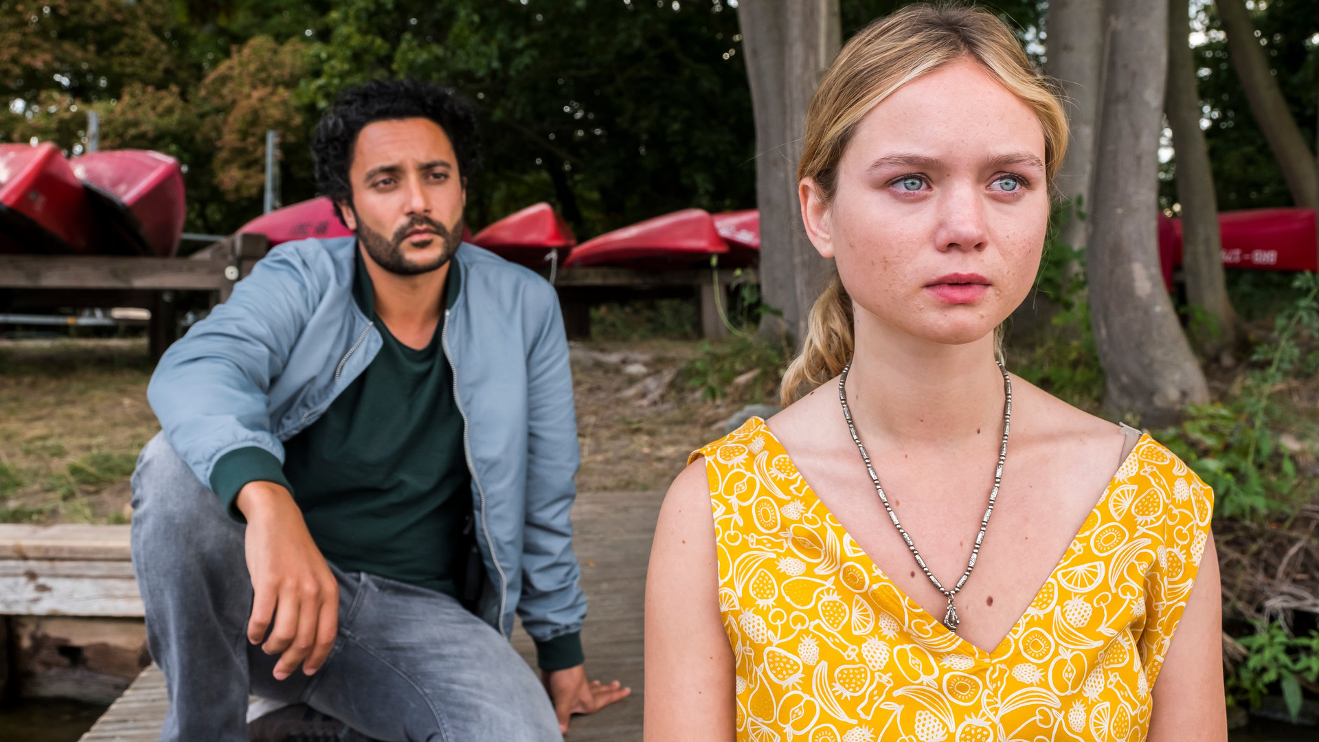 "SOKO Potsdam - Hass ist mein Hobby": Anastasia Seidel (Sofie Eifertinger) sitzt an einem Bootssteeg und blickt in die Ferne. Hinter ihr sitzt David Grünbaum (Oma El-Saeidi), der sie aufmerksam beobachtet. Im Hintergrund sieht man rote Ruderboote.