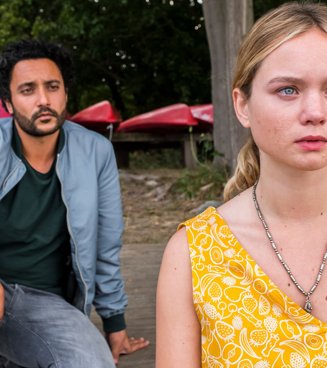 "SOKO Potsdam - Hass ist mein Hobby": Anastasia Seidel (Sofie Eifertinger) sitzt an einem Bootssteeg und blickt in die Ferne. Hinter ihr sitzt David Grünbaum (Oma El-Saeidi), der sie aufmerksam beobachtet. Im Hintergrund sieht man rote Ruderboote.