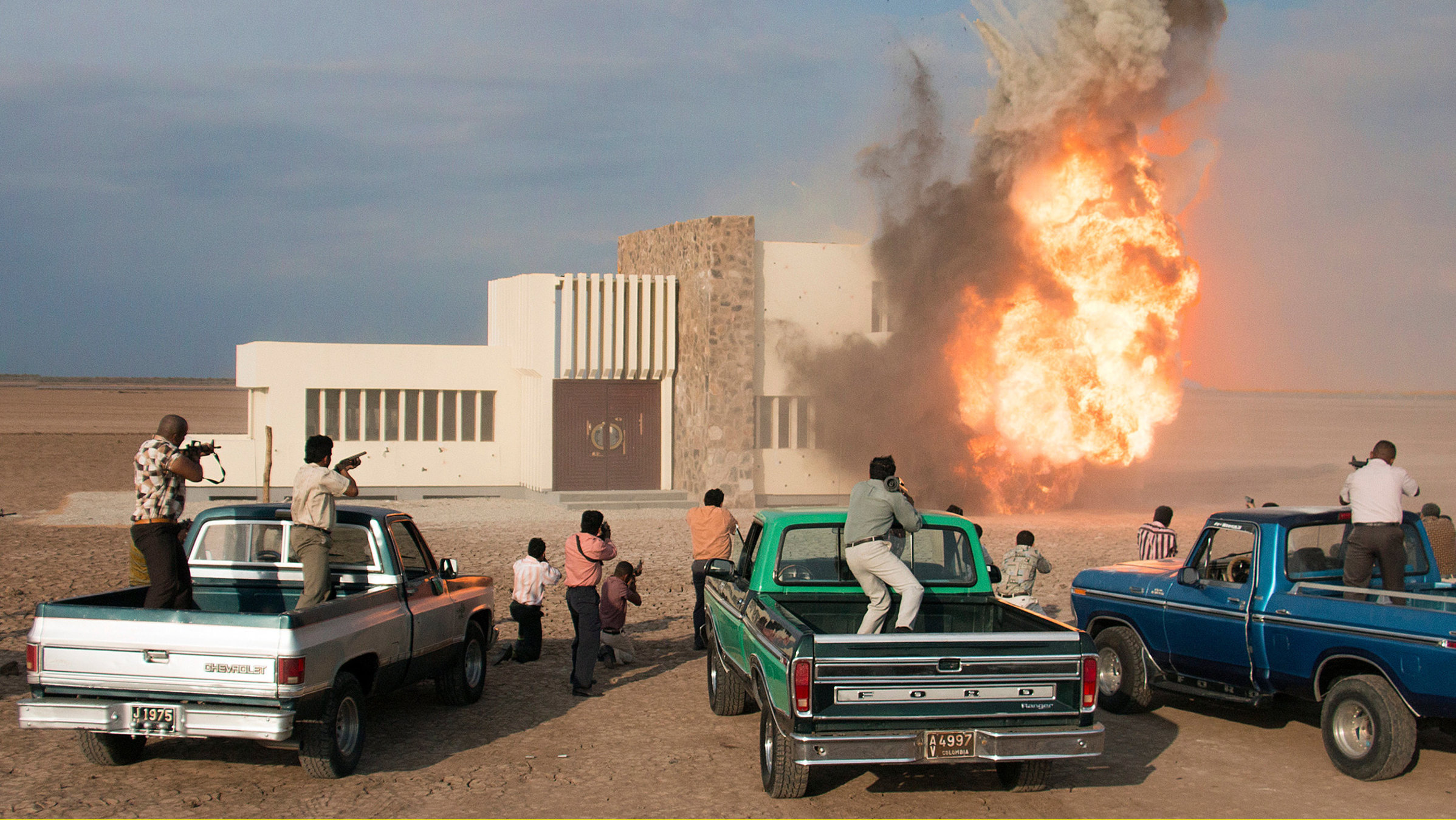 "Birds of Passage - Das grüne Gold der Wayuu": Vor einem brennendem Haus stehen mehrere Autos, auf den Autos und daneben Männer mit Schusswaffen.