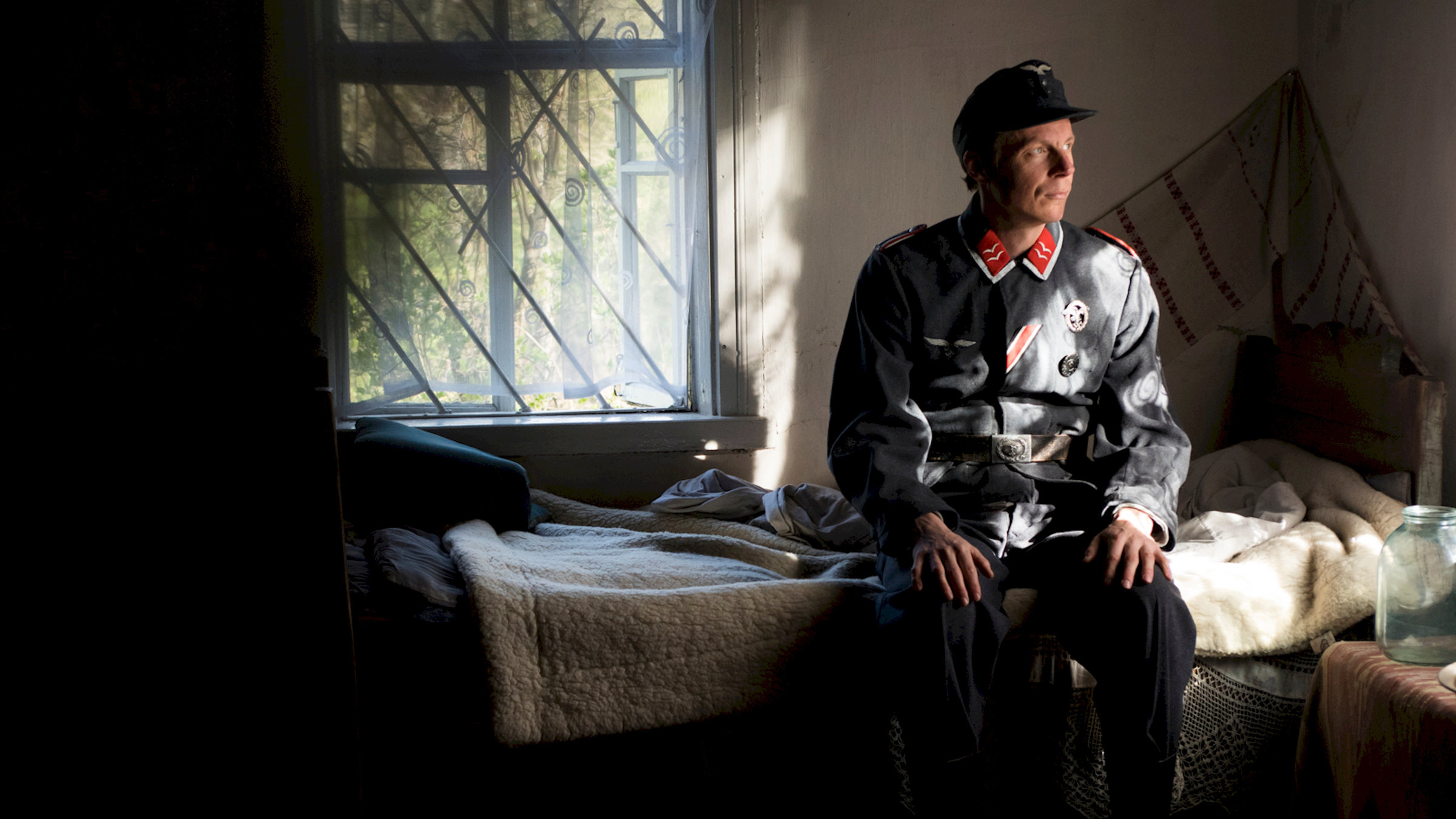 "Der Krieg in mir": Sebastian Heinzel sitzt in Wehrmachtsuniform auf einem Einzelbett im Halbdunkel und schaut nachdenklich. Im Hintergrund sieht man Bäume durch das geöffnete Fenster.